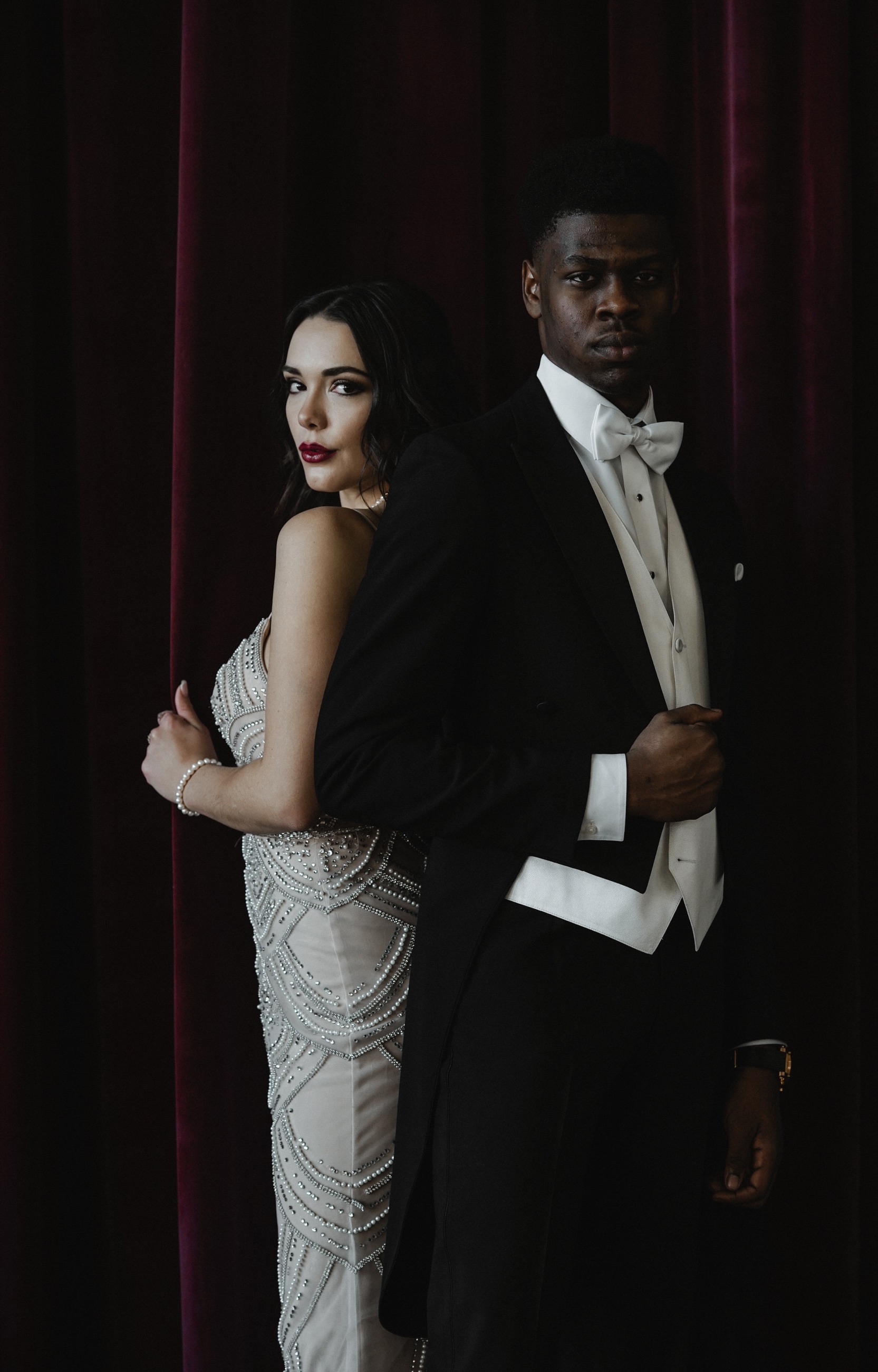 A formal couple standing back-to-back against deep red curtains