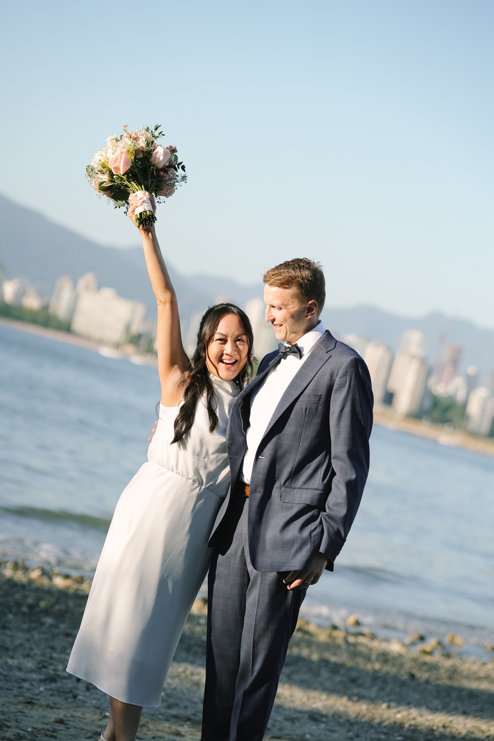 A joyful newlywed couple raising a bouquet beside the waterfront
