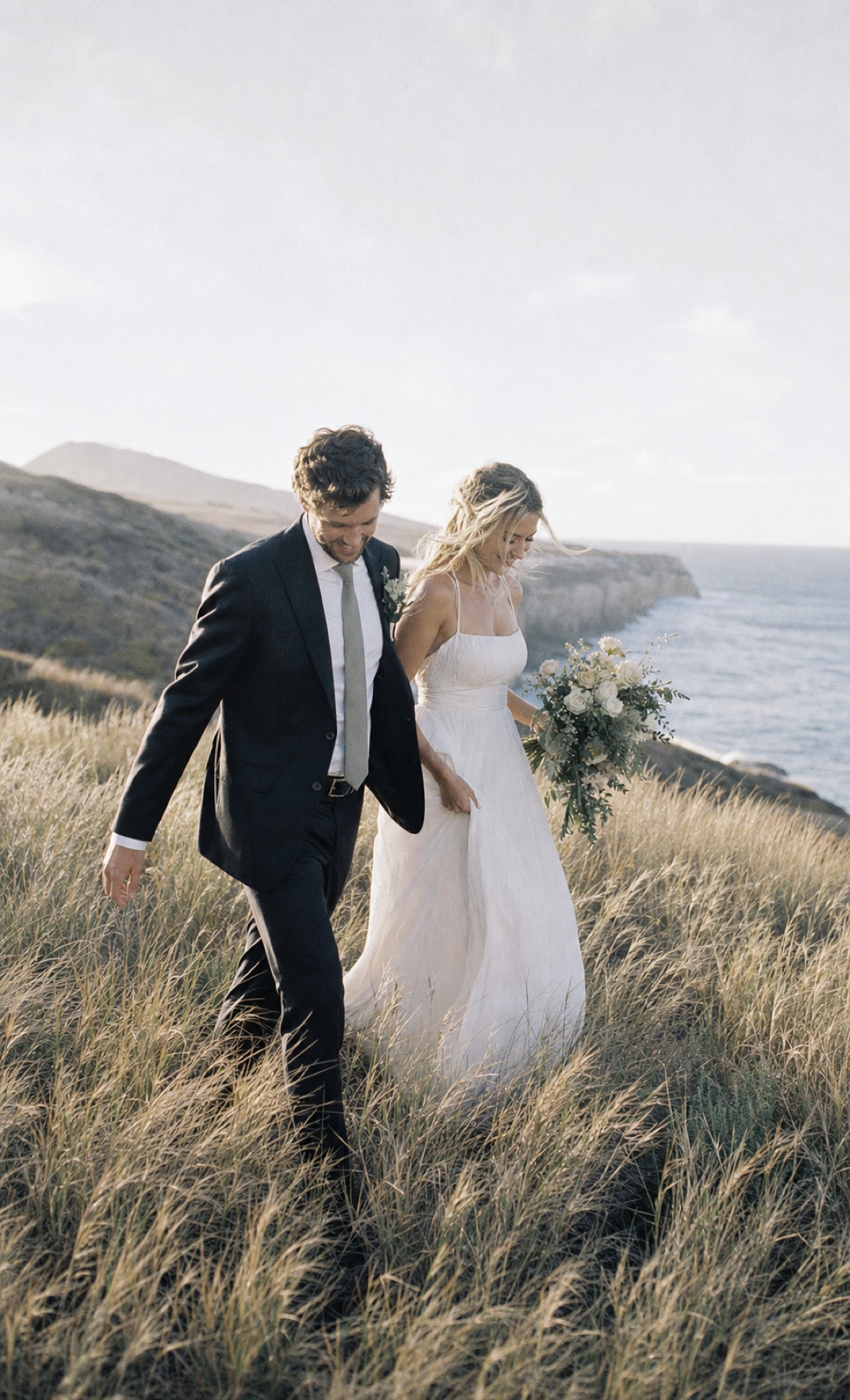 A newlywed couple walking through coastal grass